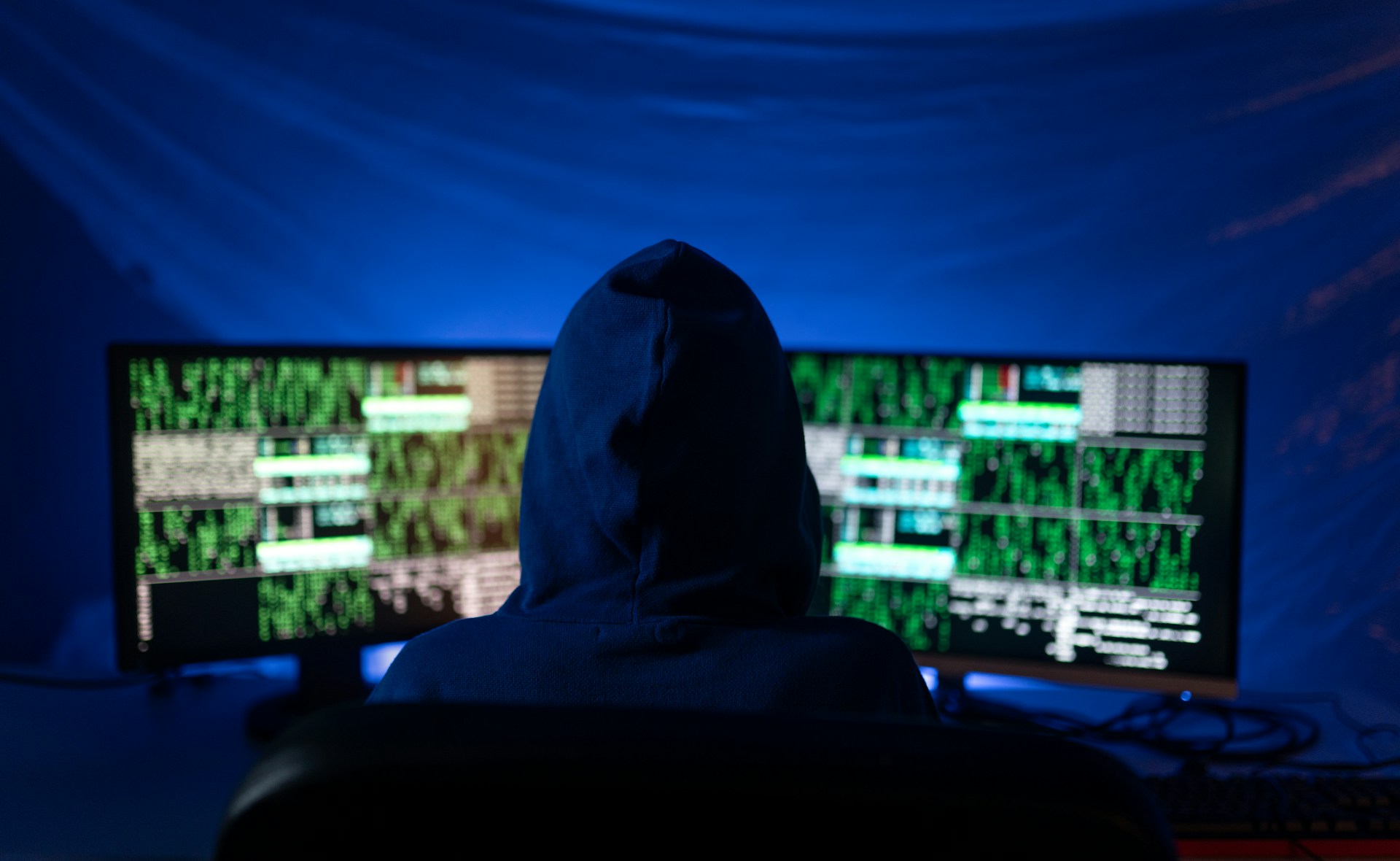 A hooded figure sits in front of a computer with hacking styled themes on the desktop computer