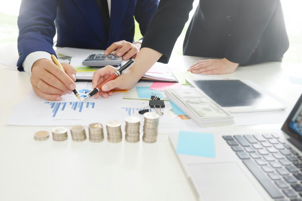 Two people in suits reviewing financial charts at a desk with stacked coins, cash, a calculator, and a laptop