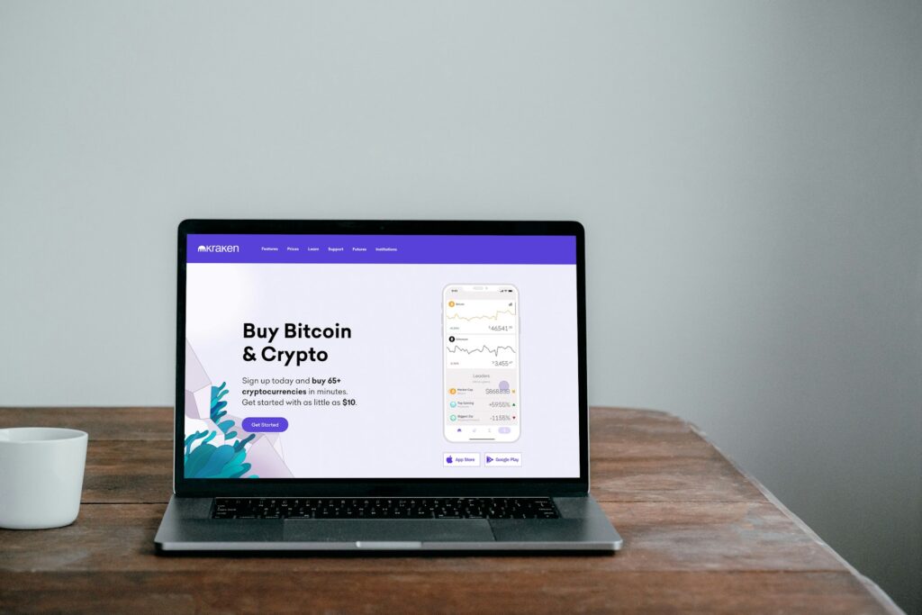 Laptop on a wooden desk displaying the kraken crypto exchange homepage with a coffee cup beside it