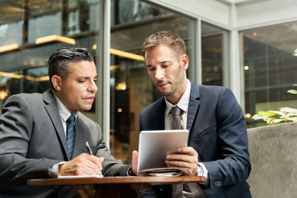 two businessmen sit on a computer outdoors talking through a business banking project