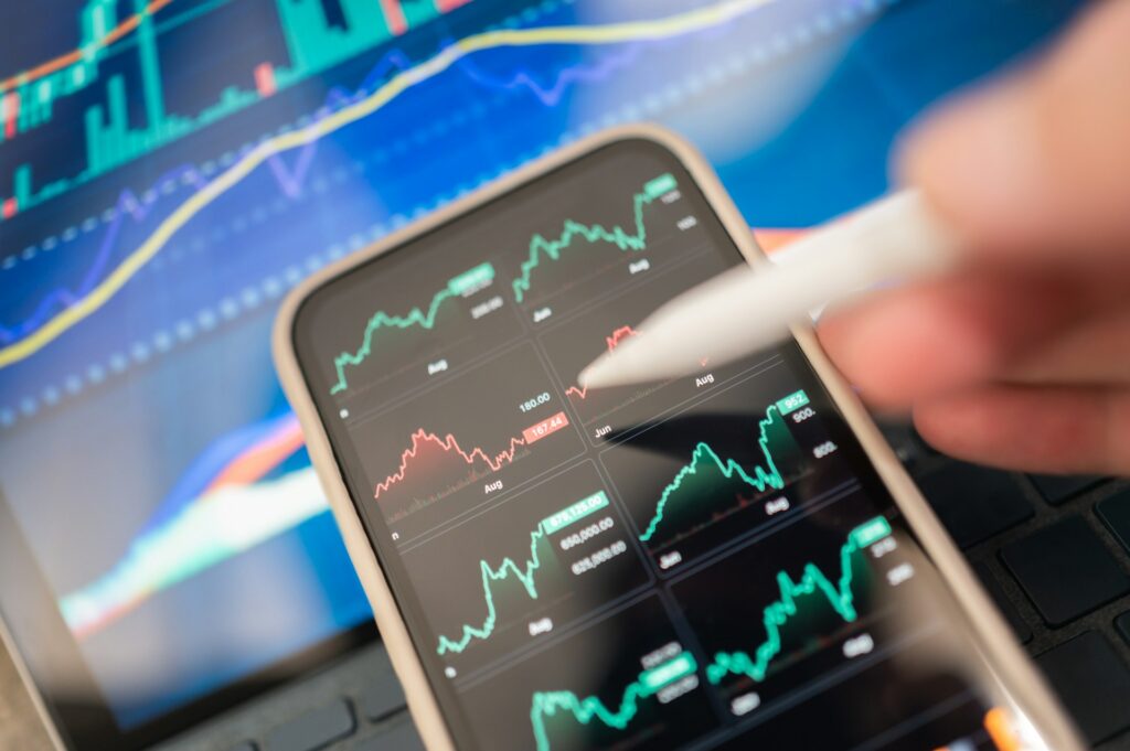 Hand holding a stylus over a phone displaying market charts, laptop behind